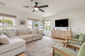 A living room with a sectional sofa, a flat-screen TV, a ceiling fan, and a large window with a view of the outdoors.