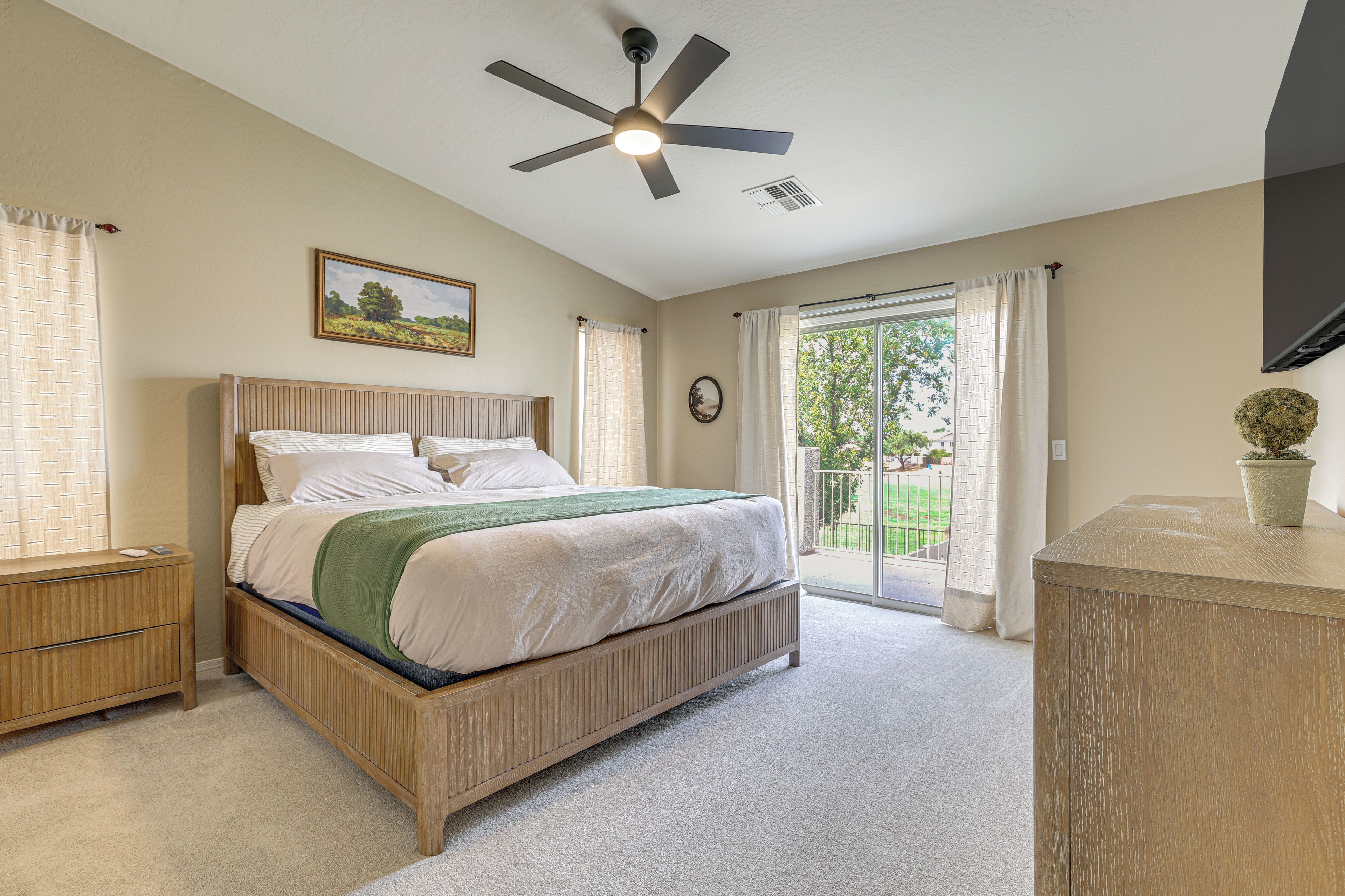 A bedroom with a bed, a ceiling fan, a window with curtains, a painting on the wall, and a potted plant.