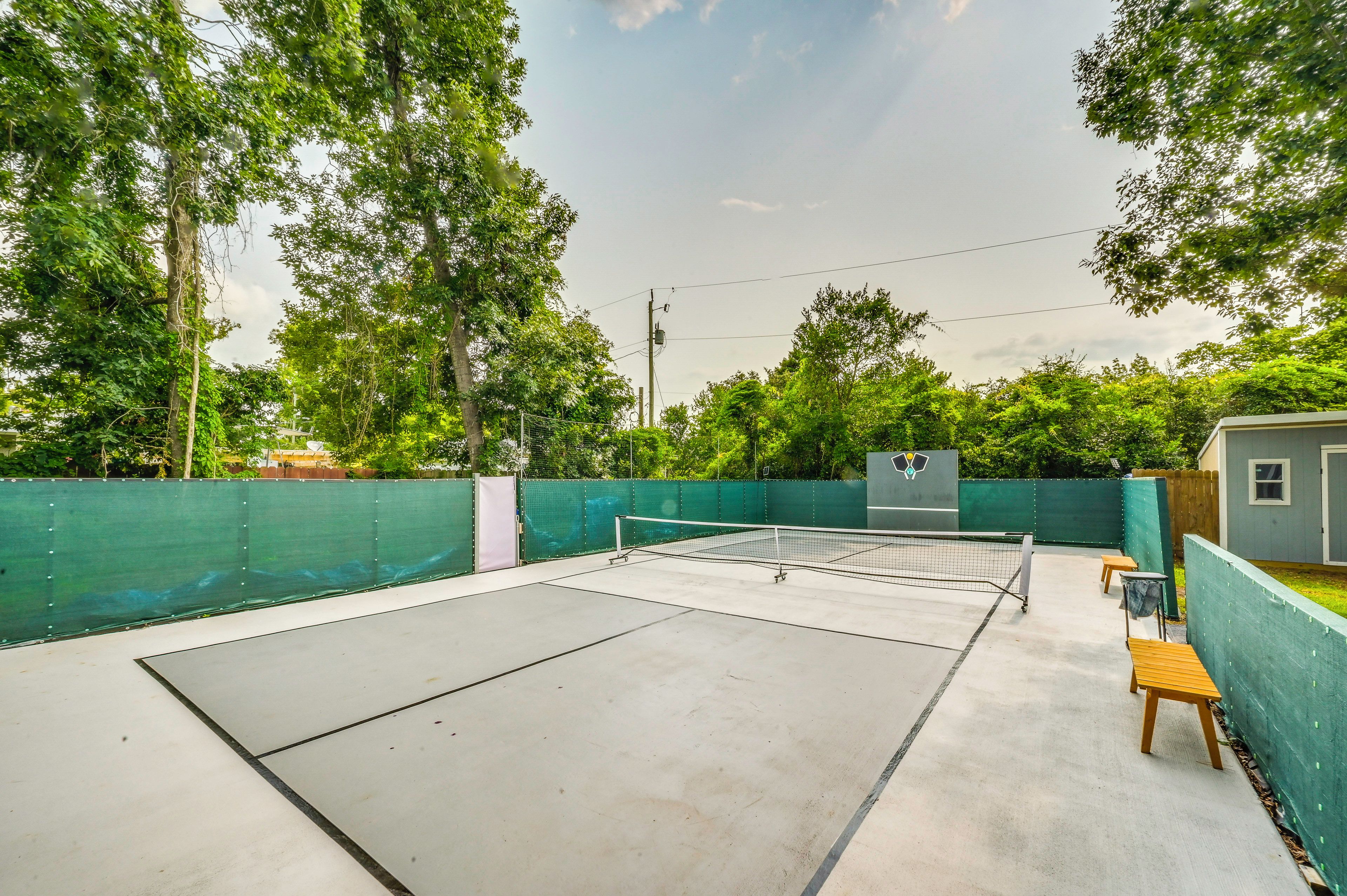 An outdoor tennis court with a net, surrounded by a green fence, trees, and a small building.