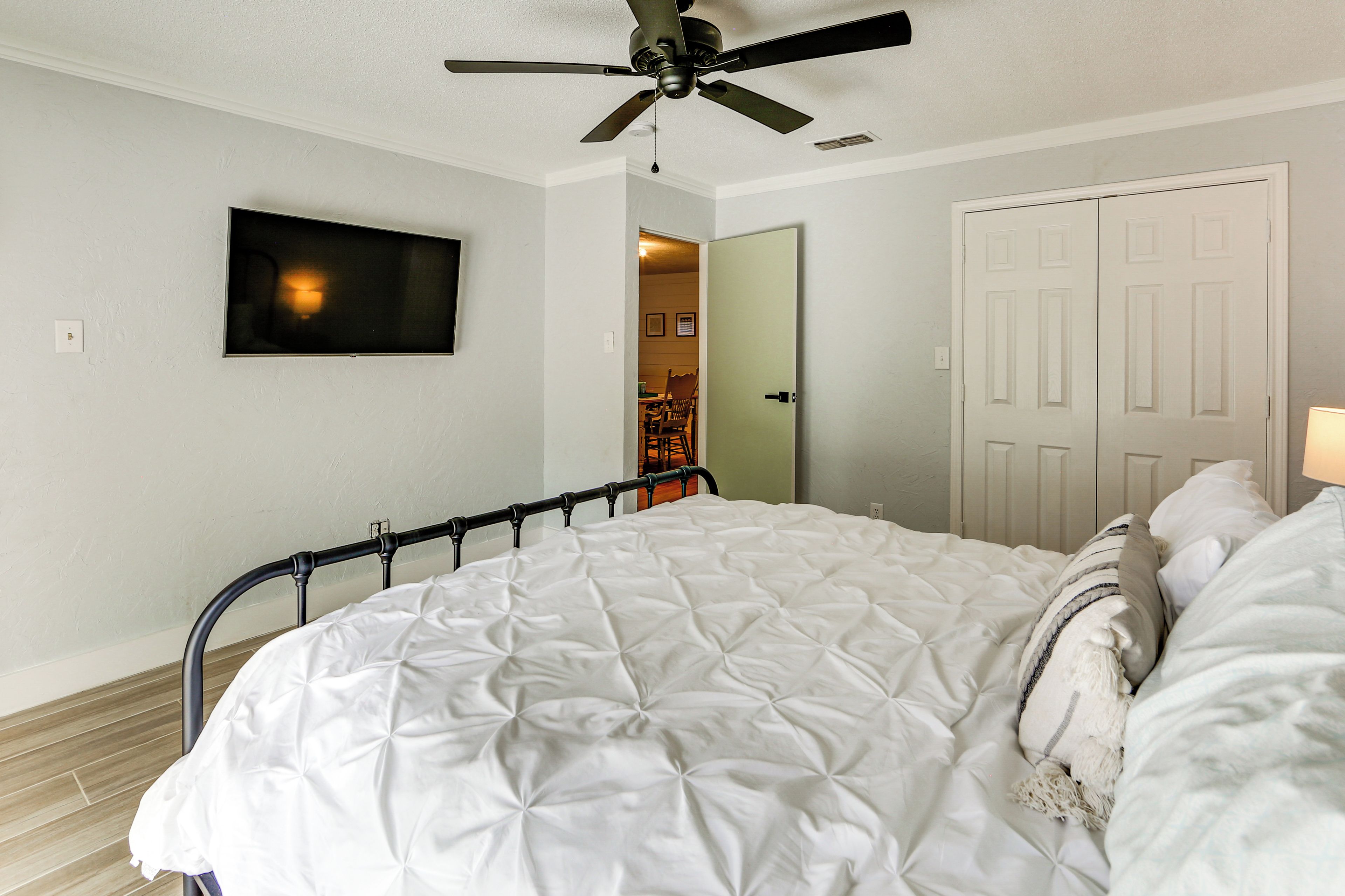 Une chambre à coucher avec un lit, un ventilateur de plafond, un téléviseur fixé au mur et une porte donnant sur une autre pièce.