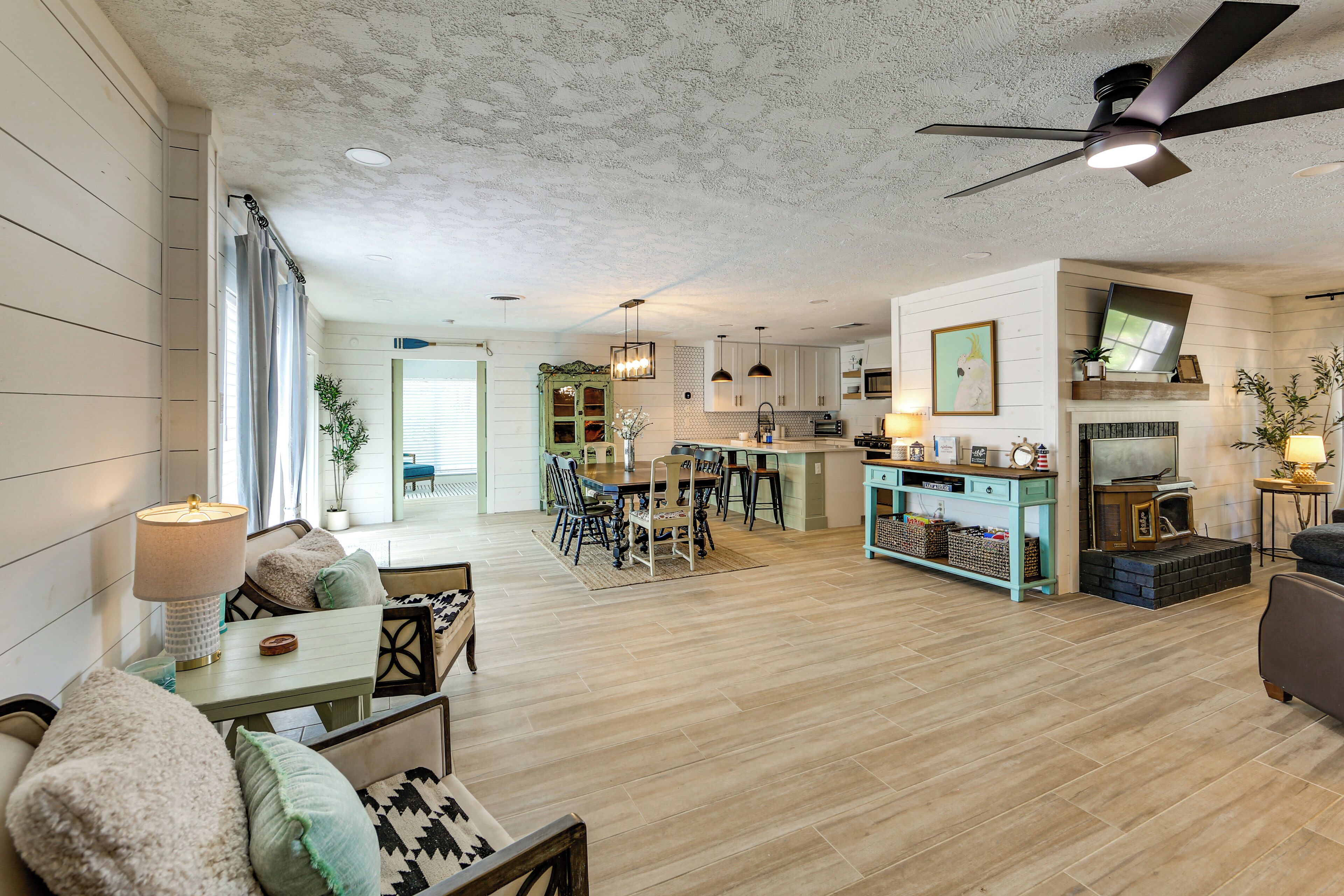 A spacious living area with a dining table, chairs, and a sofa. There is a kitchen area with modern appliances and a dining table. The room has wooden flooring and a ceiling fan.