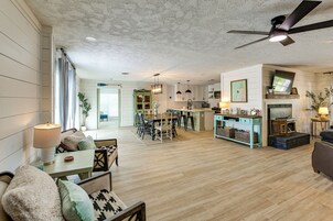 Amplia sala de estar con mesa de comedor, sillas y sofá. Hay una zona de cocina con electrodomésticos modernos y una mesa de comedor. La habitación tiene piso de madera y un ventilador de techo.