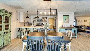 Uma sala de jantar com mesa e cadeiras de madeira, uma cozinha ao fundo, e uma sala de estar com sofá e televisão.
