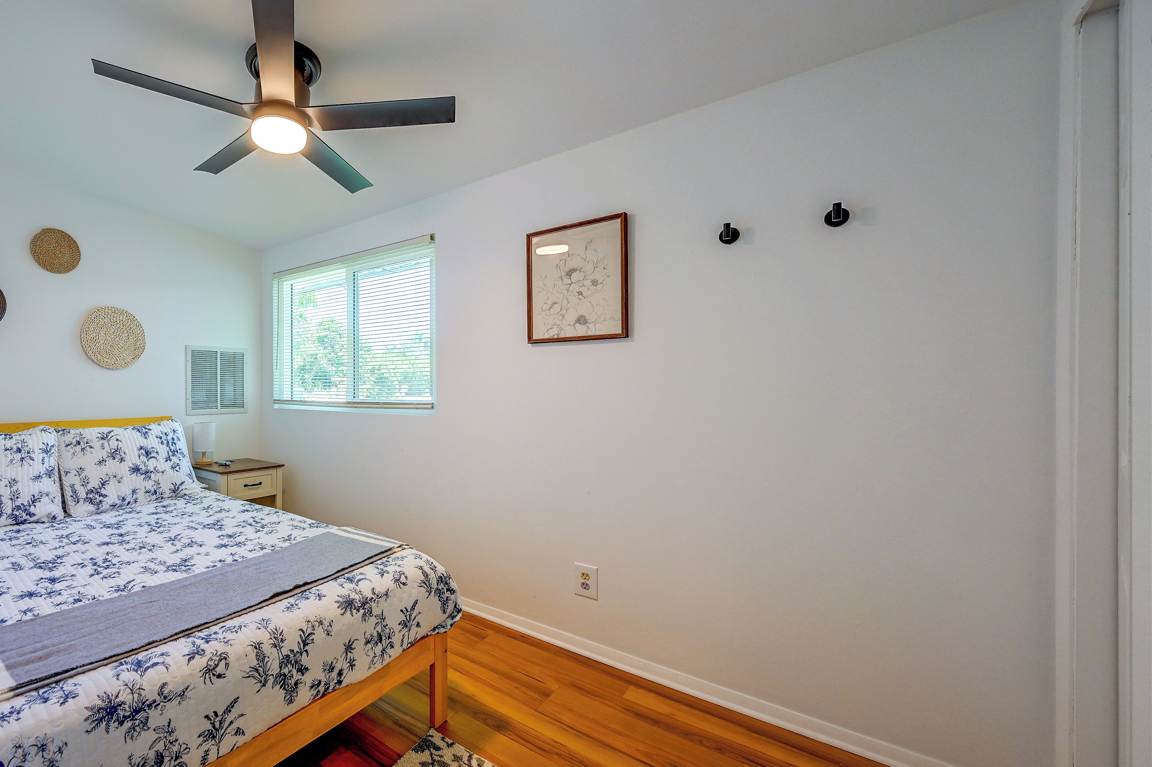 A bedroom with a bed, a ceiling fan, a window with blinds, a framed picture, and a wall socket.