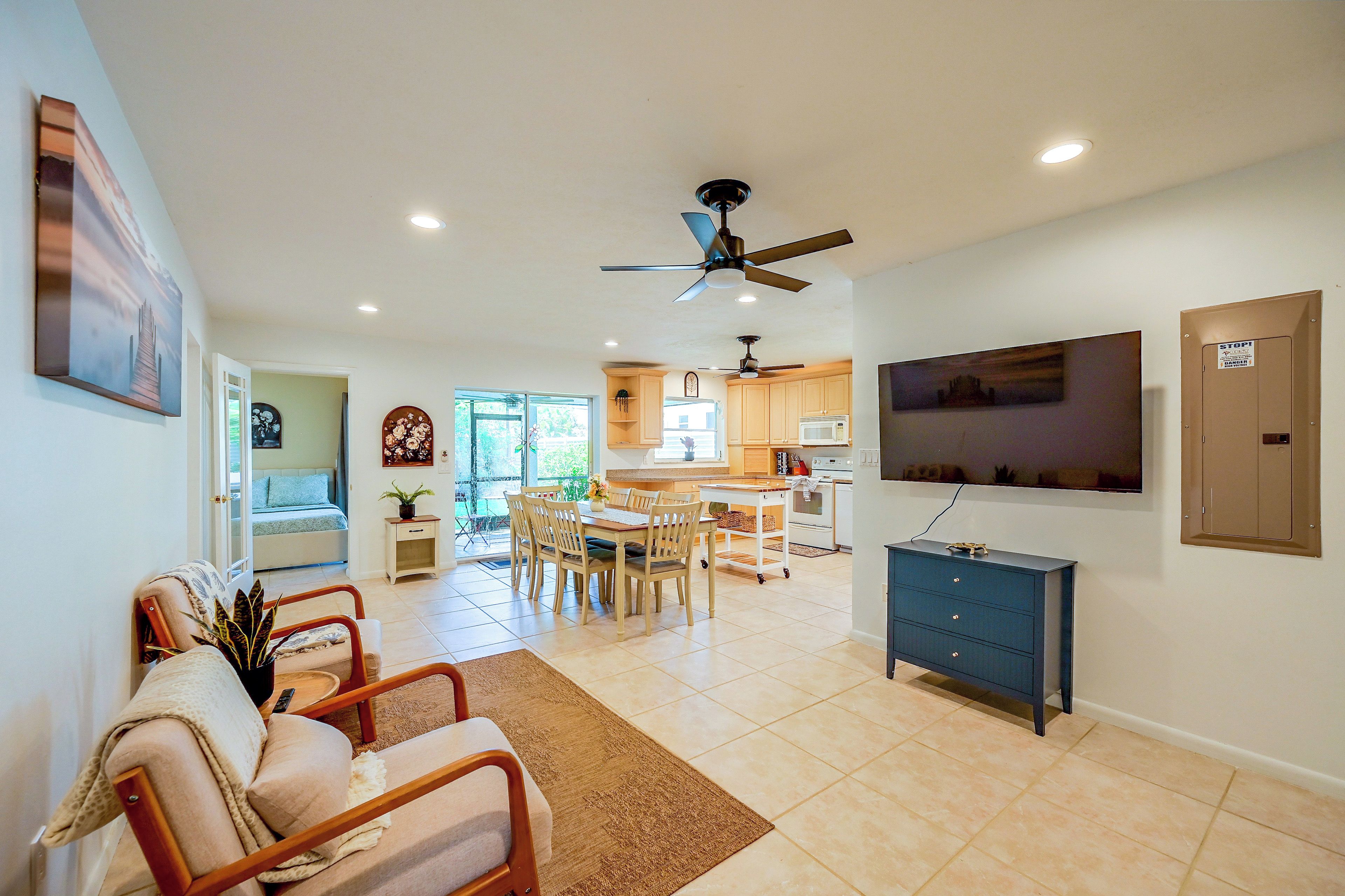 A spacious living area with a dining table, chairs, a sofa, a TV mounted on the wall, and a ceiling fan.