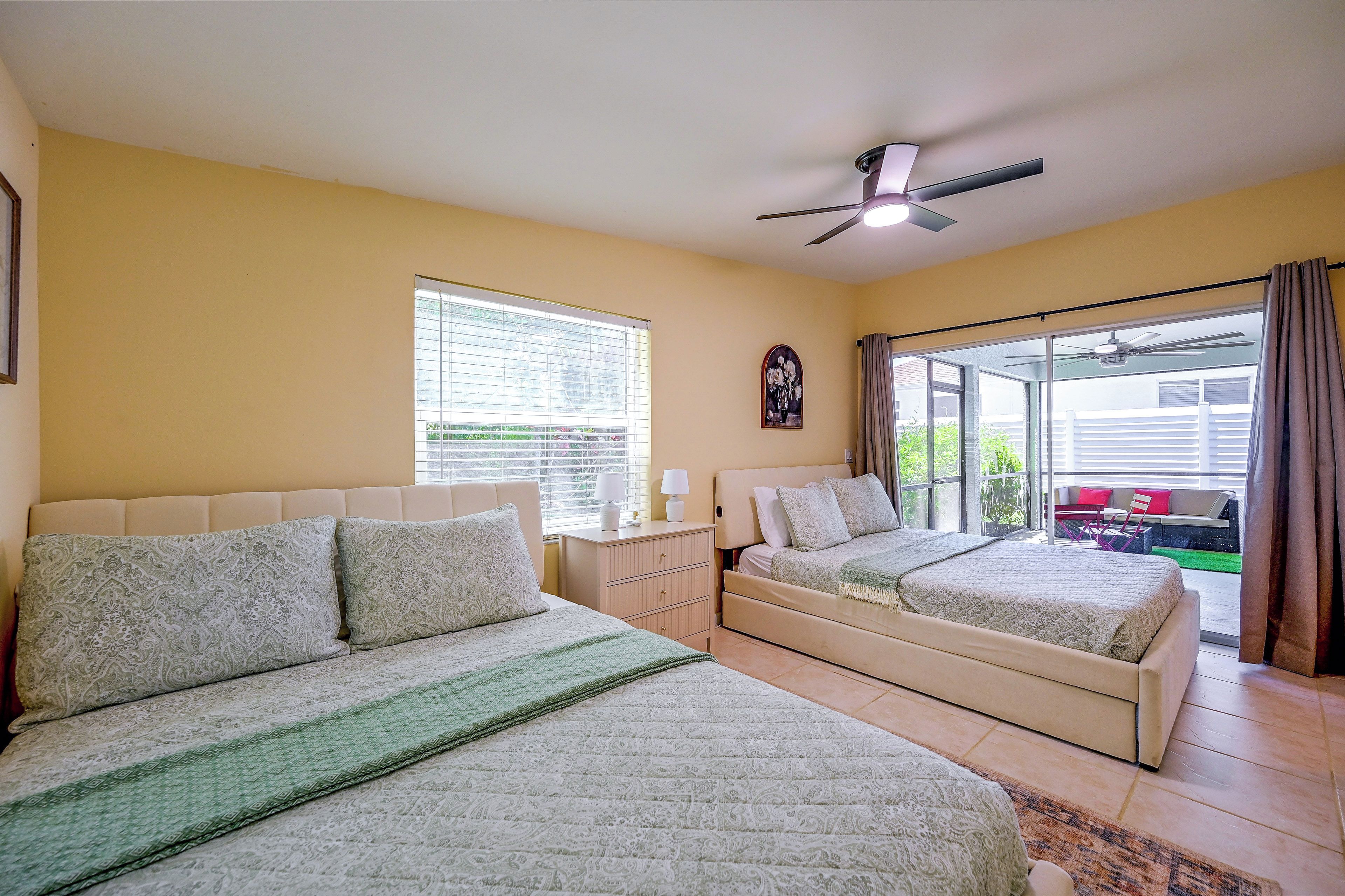 A bedroom with two beds, a ceiling fan, a window with blinds, and a view of the outside.