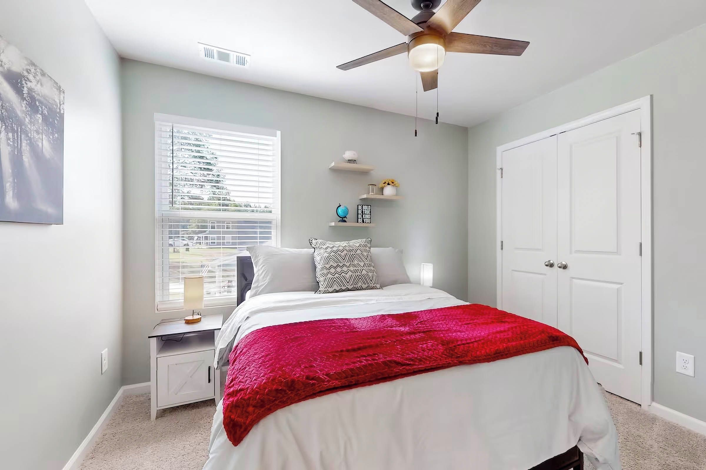 Un dormitorio con cama, mesita de noche, ventilador de techo, ventana con persianas, puerta y estante montado en la pared con objetos decorativos.