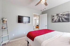 Quarto com cama, televisão, ventilador de teto, porta e prateleira fixada na parede.