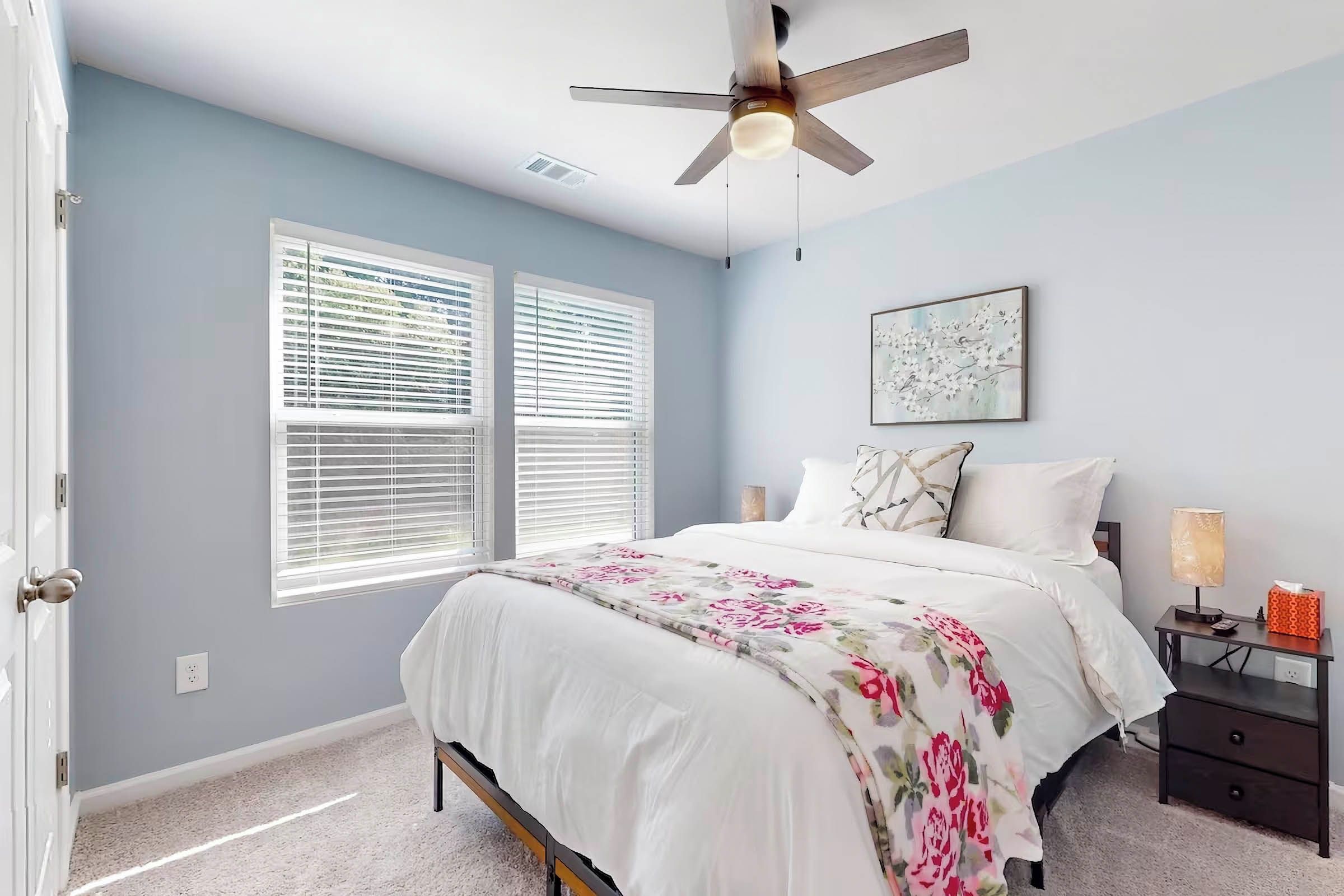 A bedroom with a bed, a nightstand, a lamp, a ceiling fan, and two windows with blinds.