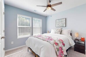 A bedroom with a bed, a nightstand, a lamp, a ceiling fan, and two windows with blinds.