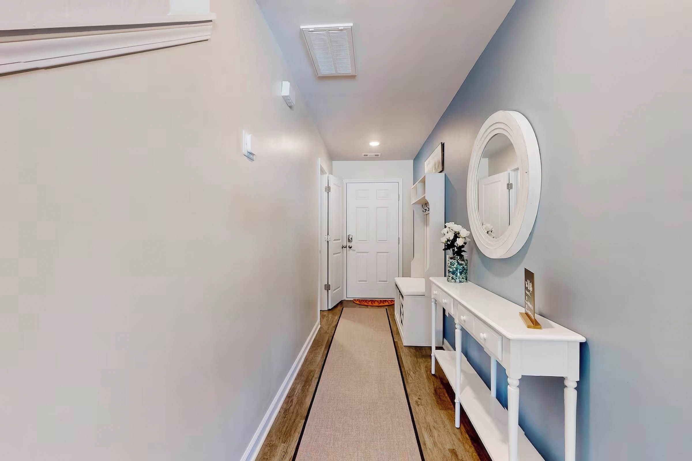 Un couloir avec une porte blanche au fond, une console blanche ornée d’un miroir et d’un vase de fleurs, et un plancher en bois.