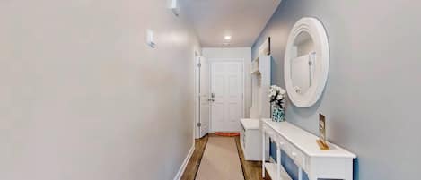 Un pasillo con una puerta blanca al final, una consola blanca con espejo y un florero, y suelo de madera.