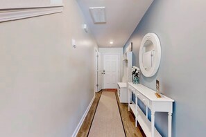A hallway with a white door at the end, a white console table with a mirror and a vase of flowers, and a wooden floor.