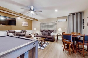 Ein modernes Wohnzimmer mit Billardtisch, Ledersofas, einem Esstisch mit Stühlen und einem Deckenventilator.