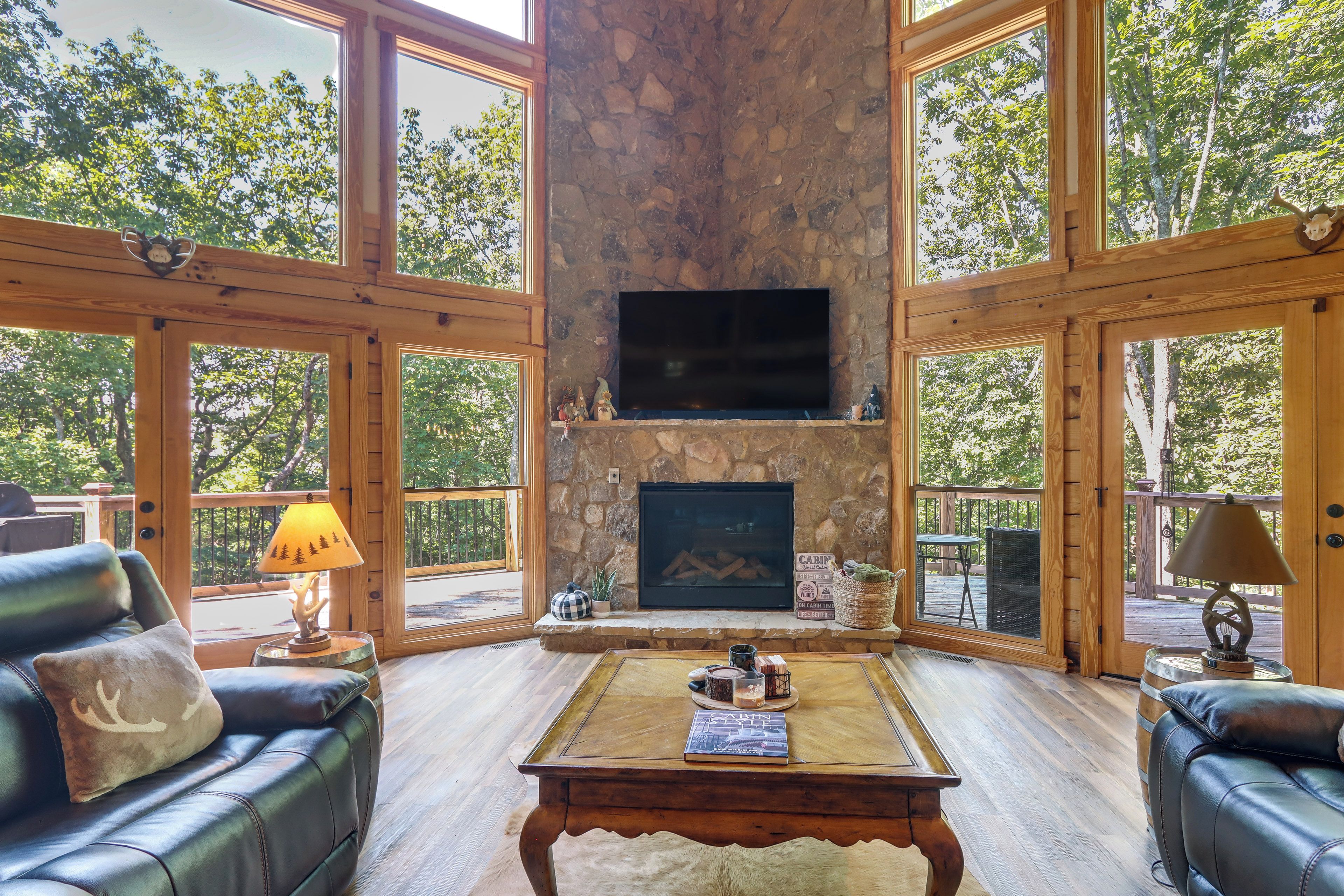 A spacious living room with a stone fireplace, leather sofas, a coffee table, and large windows offering a view of the surrounding trees.