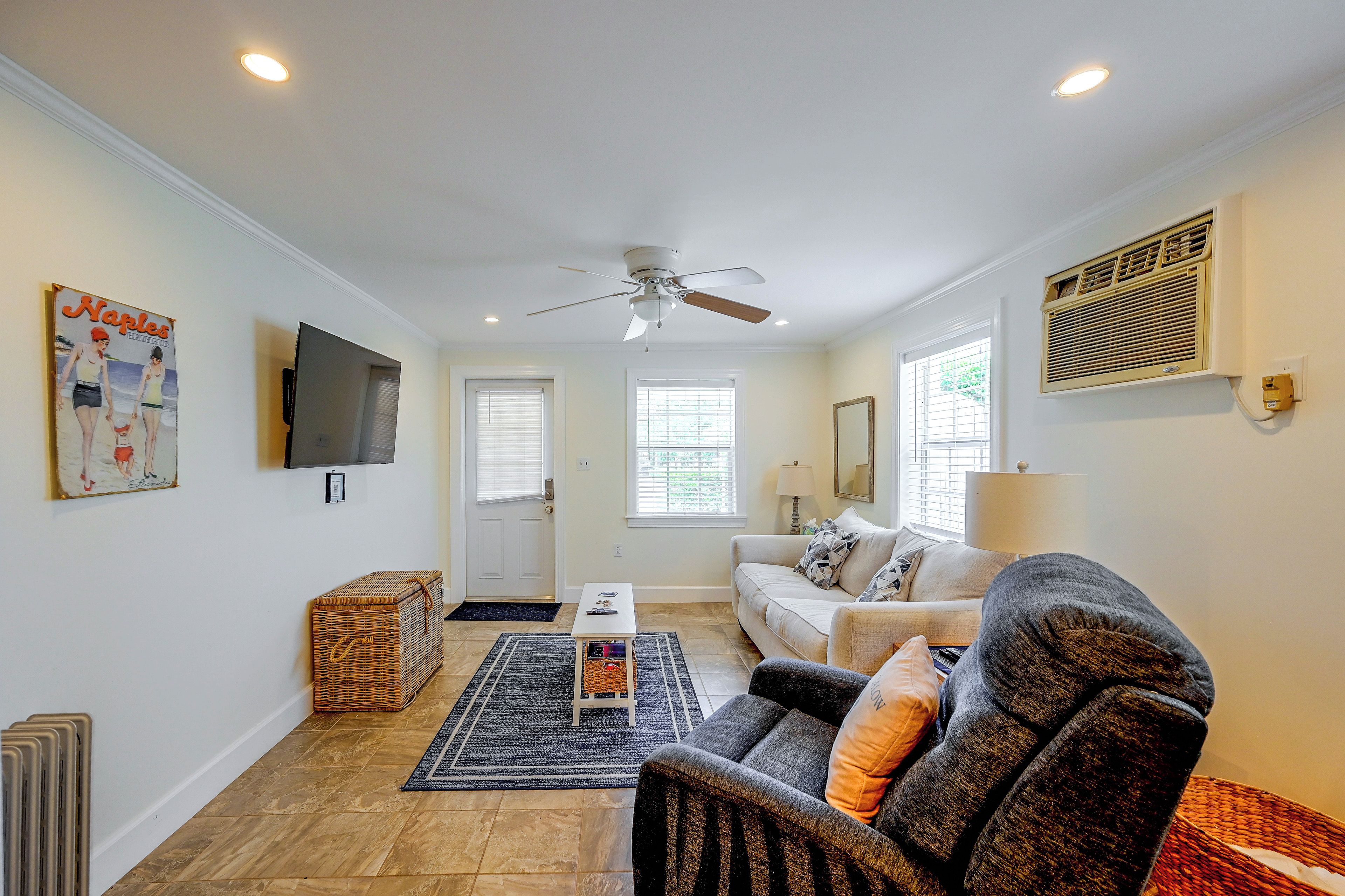 A living room with a ceiling fan, a flat-screen TV mounted on the wall, a sofa with cushions, a coffee table, and a wicker basket.