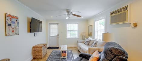 A living room with a ceiling fan, a flat-screen TV mounted on the wall, a sofa with cushions, a coffee table, and a wicker basket.