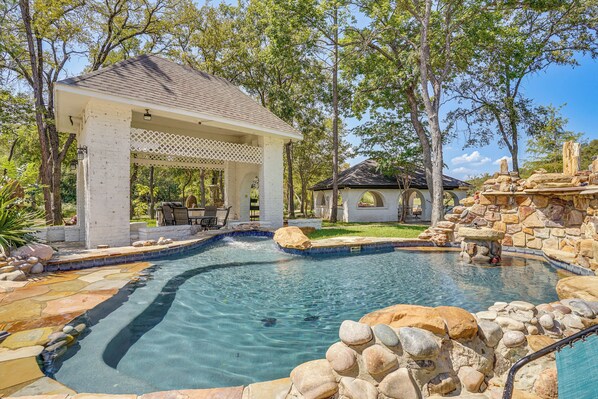 A swimming pool with a stone border, a gazebo, and a small building in a landscaped area.