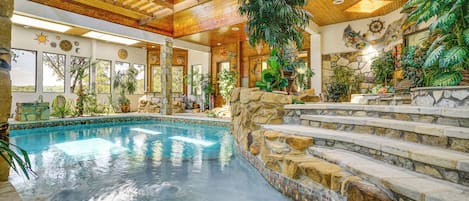 Una piscina con escalones de piedra que llevan a una terraza de madera, rodeada de plantas y elementos decorativos.