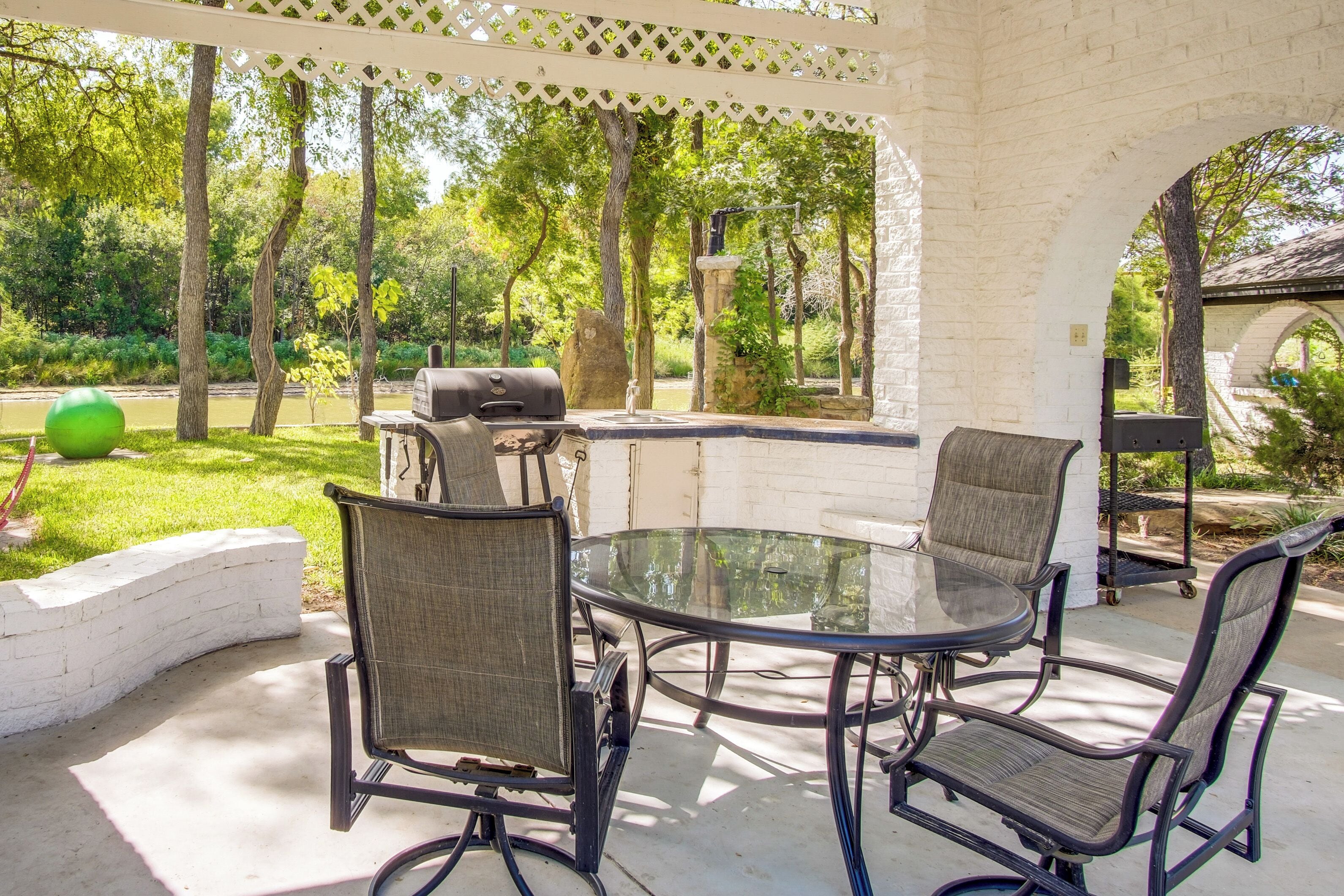 Une terrasse extérieure avec une table en verre et quatre chaises, un barbecue et un mur en briques blanches.