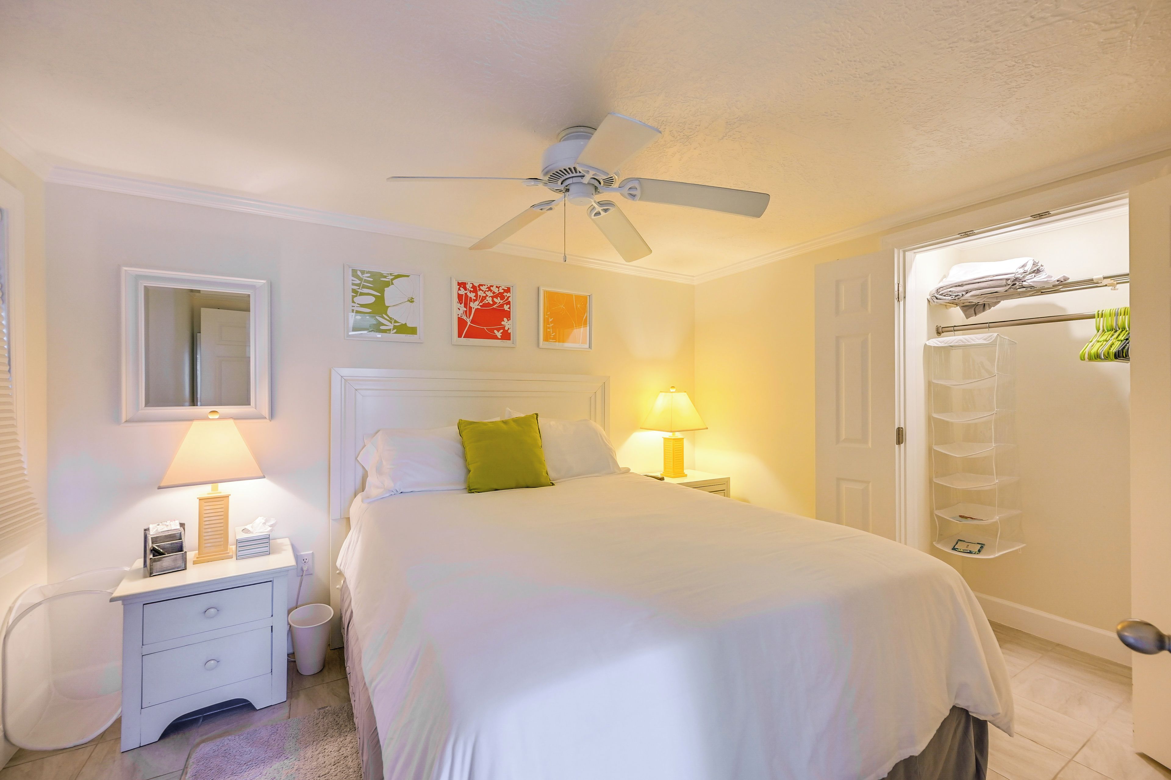 Une chambre d’hôtel comprenant un lit, des lampes de chevet, un ventilateur de plafond, une porte avec un porte-serviettes et un mur orné d’œuvres d’art encadrées.