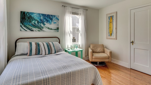 A bedroom with a bed, a chair, a window with curtains, a ceiling fan, and a door.