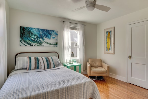 A bedroom with a bed, a chair, a window with curtains, a ceiling fan, and a door.