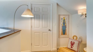 A hallway with a white door, a framed surfboard artwork, a wall-mounted lamp, and a woven pouf with a lobster design.
