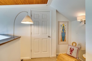 A hallway with a white door, a framed surfboard artwork, a wall-mounted lamp, and a woven pouf with a lobster design.