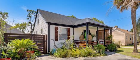 A single-story house with a covered porch, a fenced yard, and various potted plants.