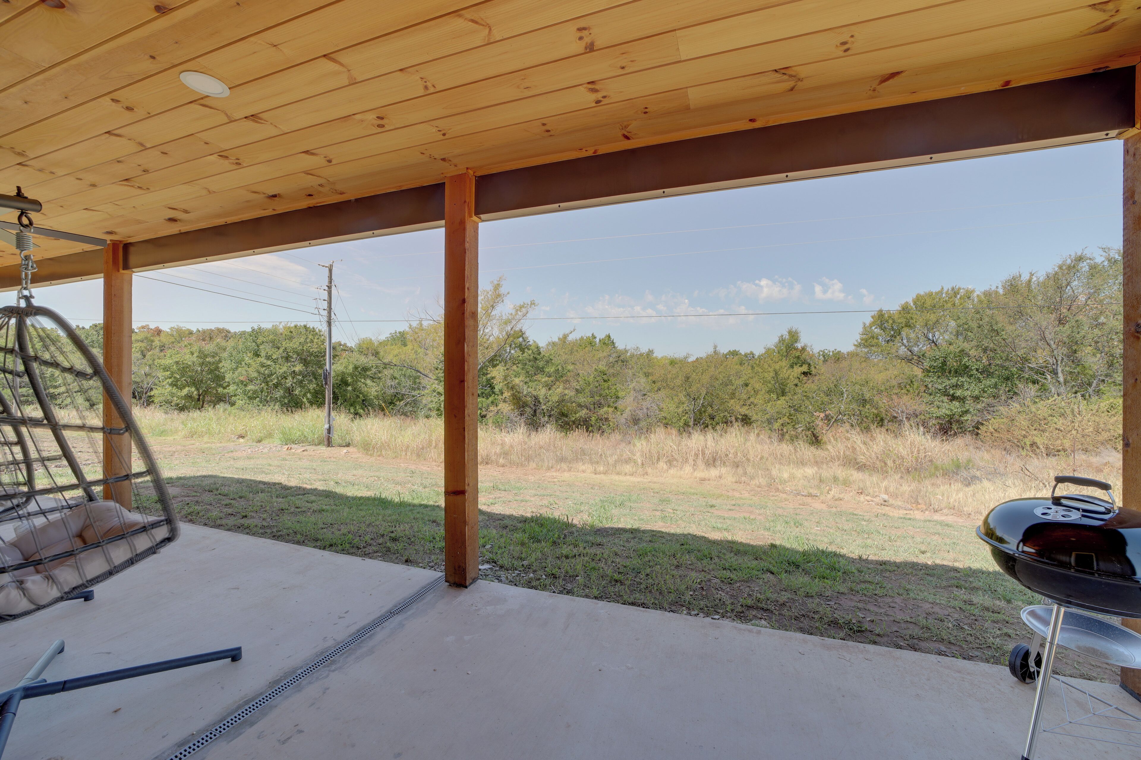 A covered outdoor area with a hanging chair, a barbecue grill, and a view of a grassy field with trees.