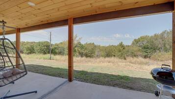 A covered outdoor area with a hanging chair, a barbecue grill, and a view of a grassy field with trees.