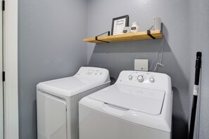 A laundry room with two front-loading washing machines and a shelf above them holding a laundry schedule, a jar of laundry detergent, and a jar of dryer sheets.