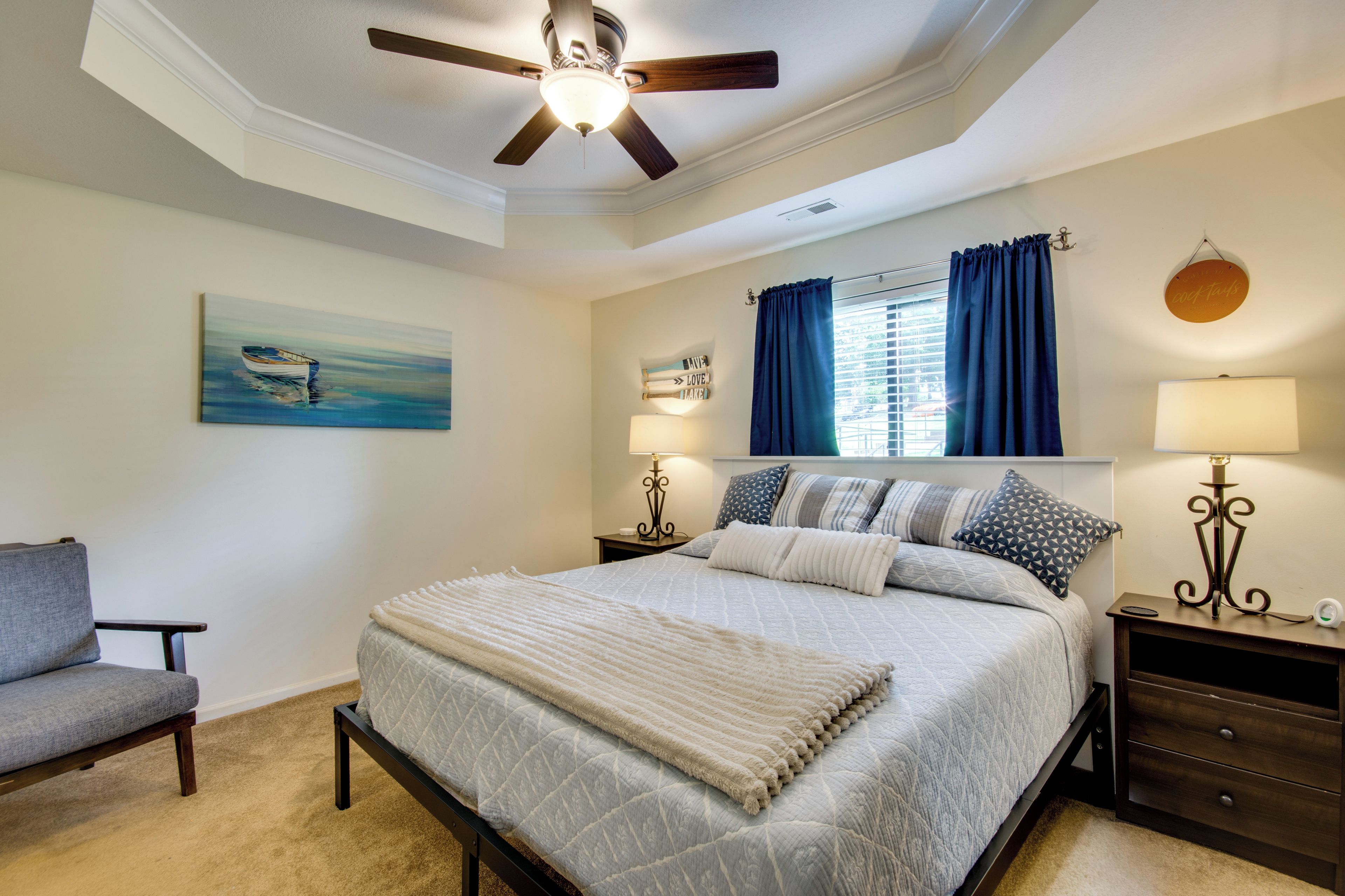 A bedroom with a bed, a chair, a ceiling fan, a painting, a window with curtains, and a bedside table with a lamp.