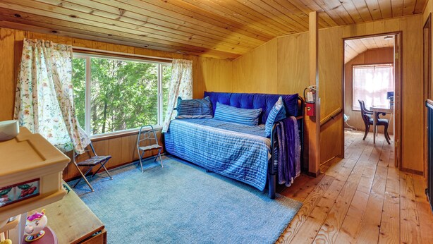 A wooden cabin interior with a bed, a small table, a chair, and a window with curtains.