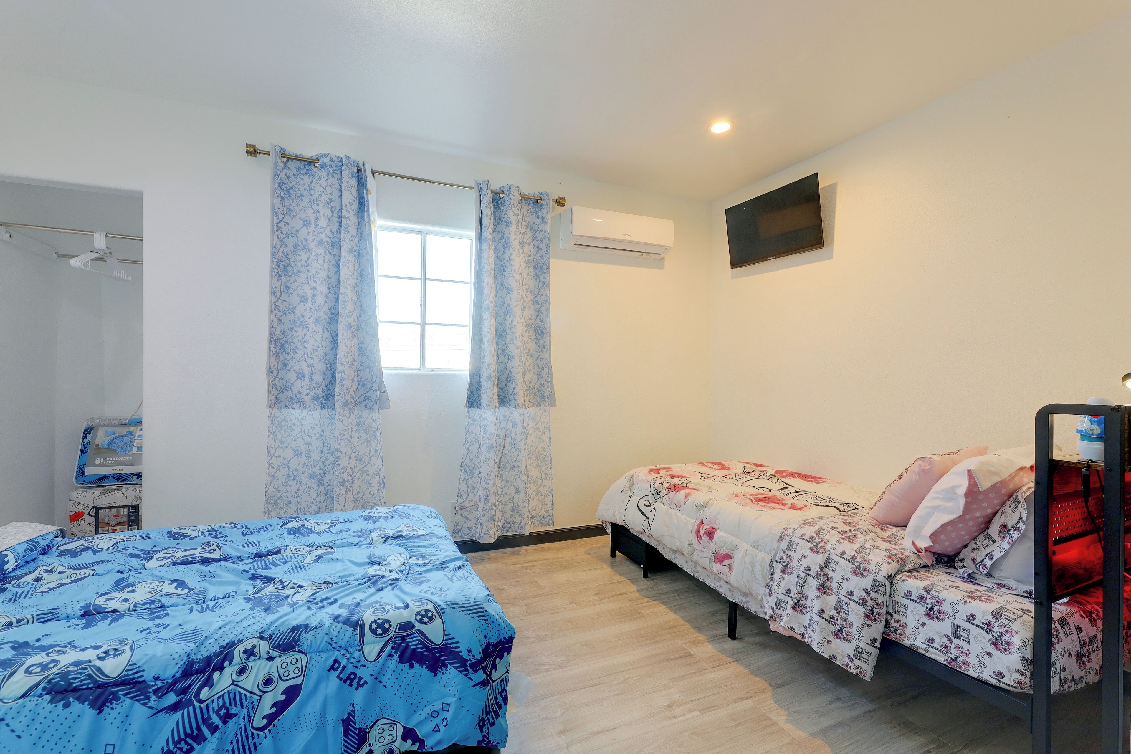 A bedroom with two beds, a window with curtains, an air conditioning unit, and a television on the wall.