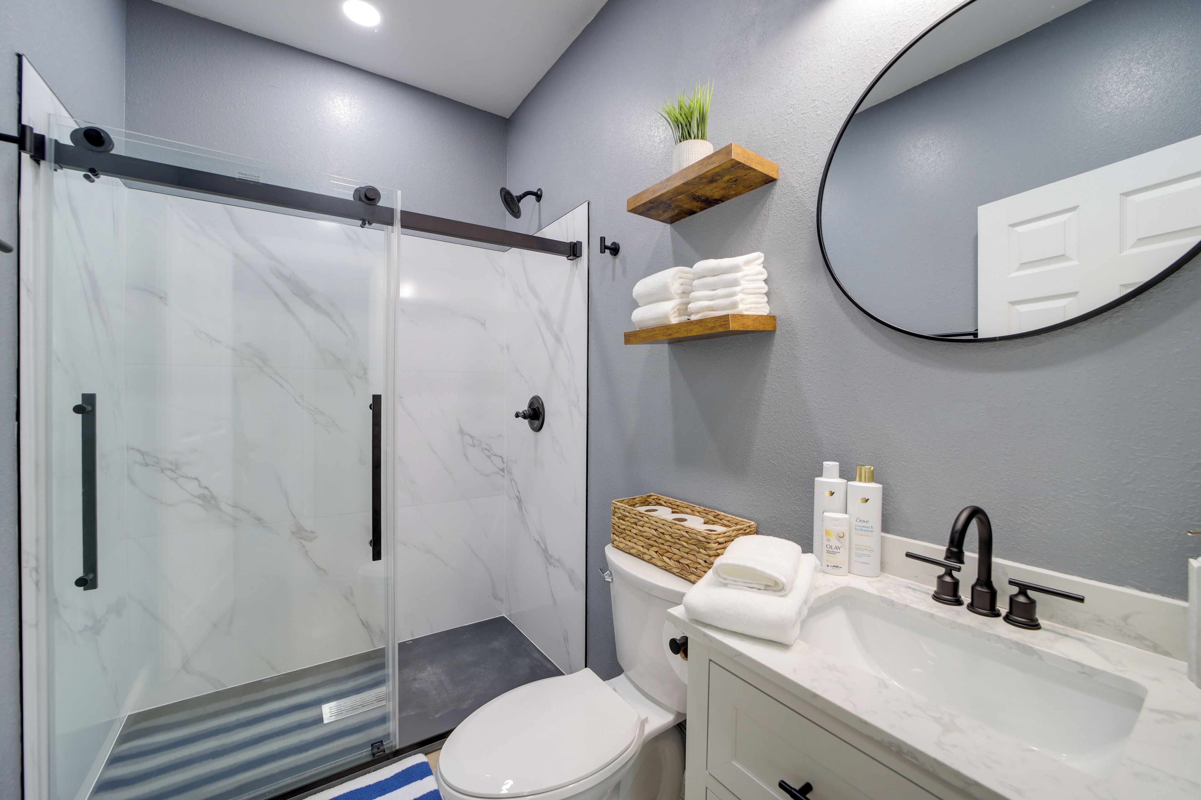 A bathroom with a glass shower enclosure, a toilet, a sink with a mirror, and a shelf with towels and toiletries.