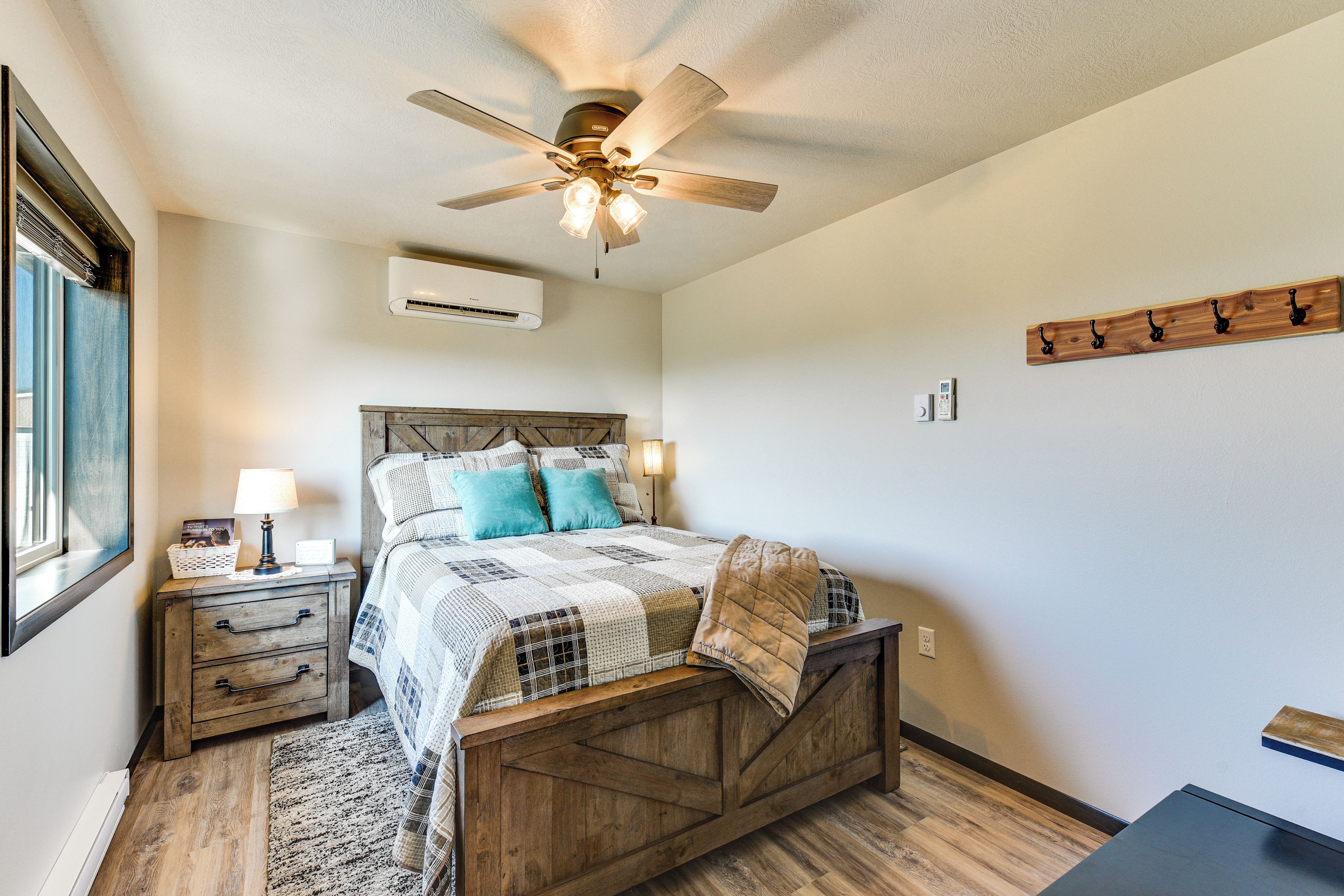 A bedroom with a wooden bed, a ceiling fan, an air conditioning unit, a bedside table with a lamp, and a wall-mounted coat rack.