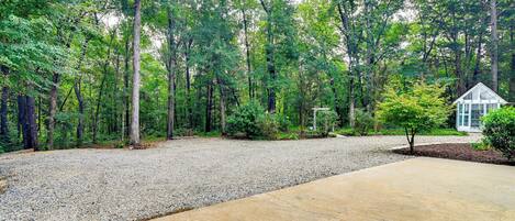 A gravel driveway leading to a house with a small garden area, surrounded by a dense forest.