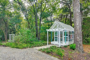 A small, white gazebo-like structure with a glass enclosure, surrounded by a forested area with a gravel path leading to it.