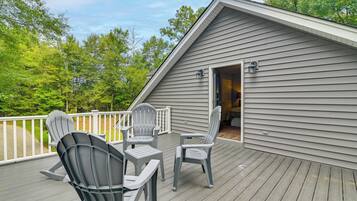 A deck with Adirondack chairs and a small table, a wooden deck, and a building with a door leading to an interior space.