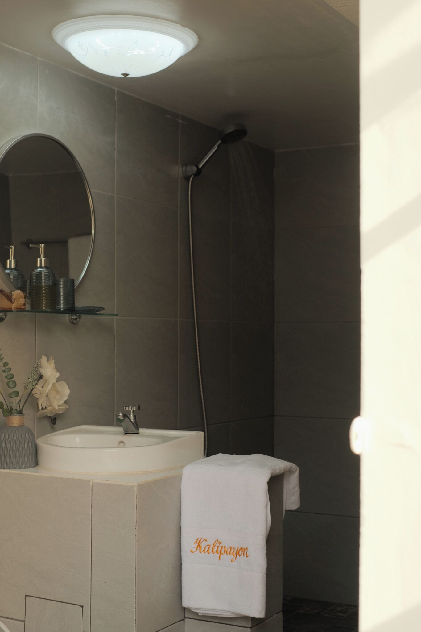 Een badkamer met een witte wastafel, een spiegel, een handdoek met de borduring 'Kalipayon' en een douchekop.