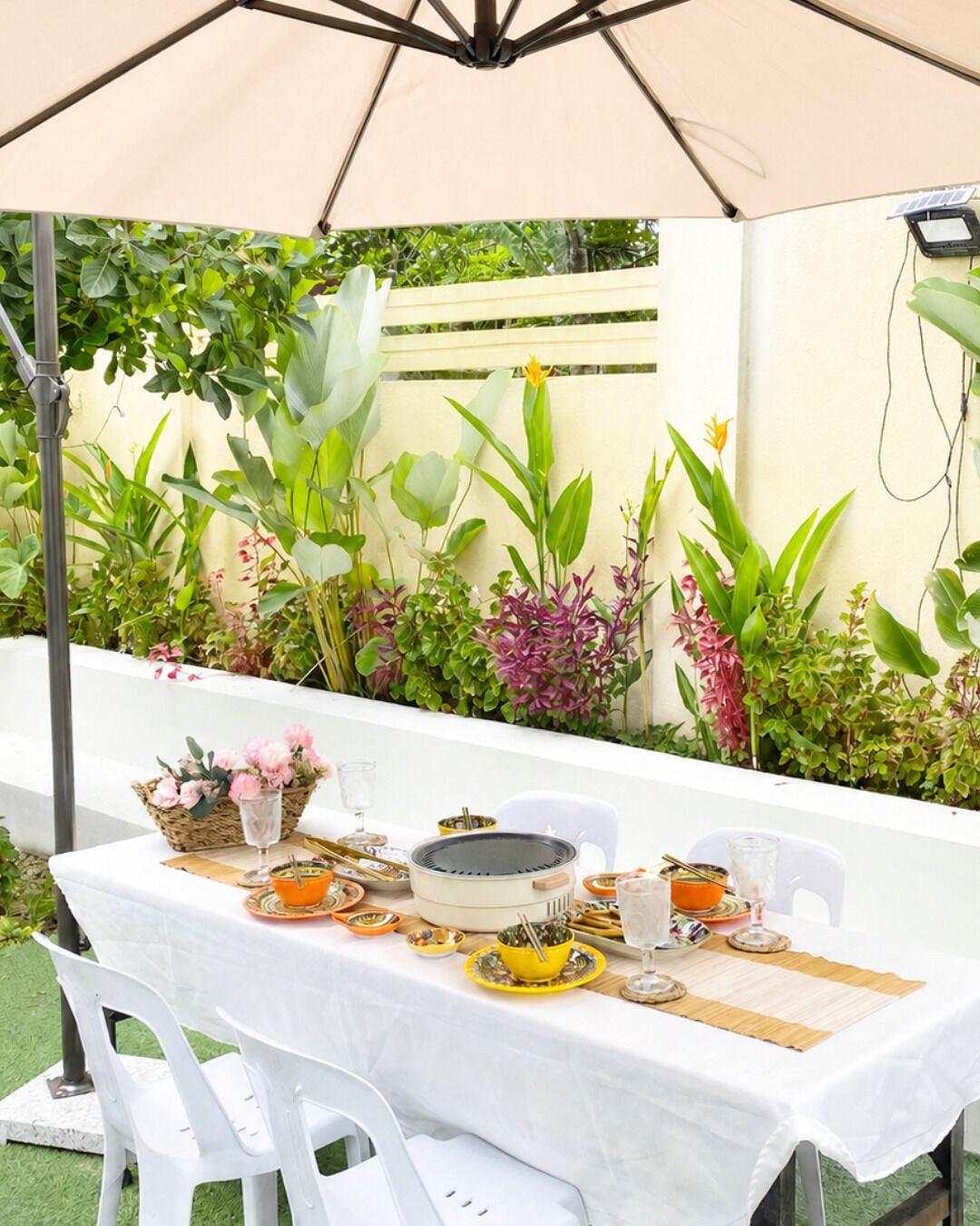Un espace de restauration en plein air avec une table dressée pour deux personnes, entouré d’une végétation luxuriante et protégé par un parasol beige.