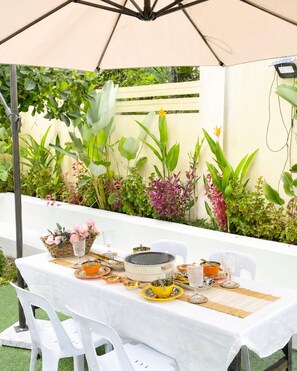Un área de comedor al aire libre con una mesa lista para dos personas, rodeada de abundante vegetación y un paraguas beige.