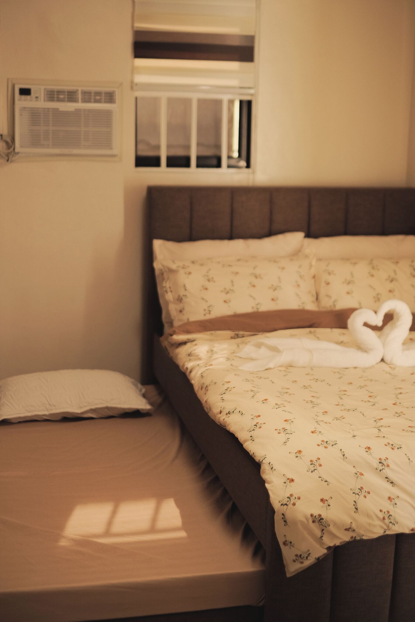 Un lit avec une couvre-lit à motifs floraux et deux oreillers, une table de chevet avec une lampe et une fenêtre avec des stores.