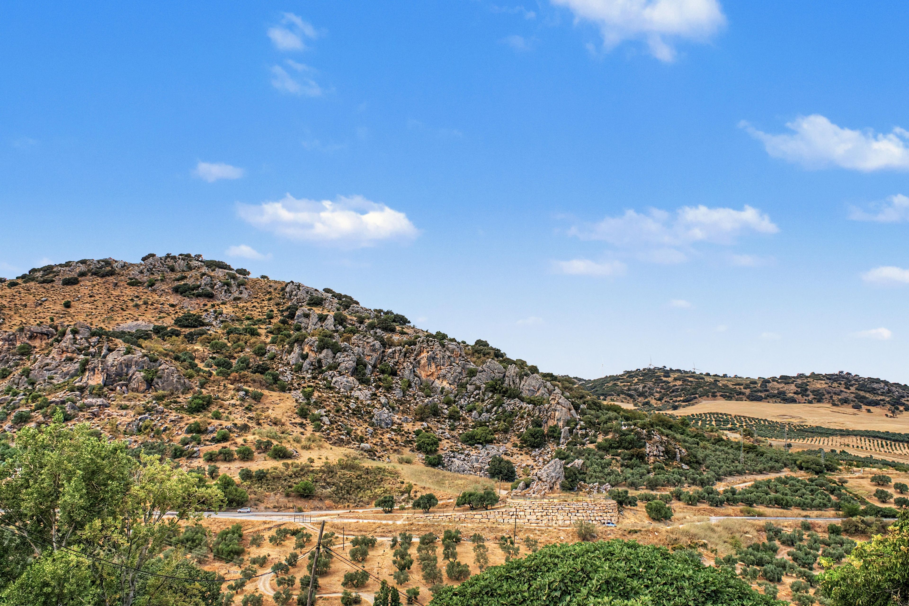 Paesaggio montuoso con cielo sereno, una strada e vegetazione sparsa.