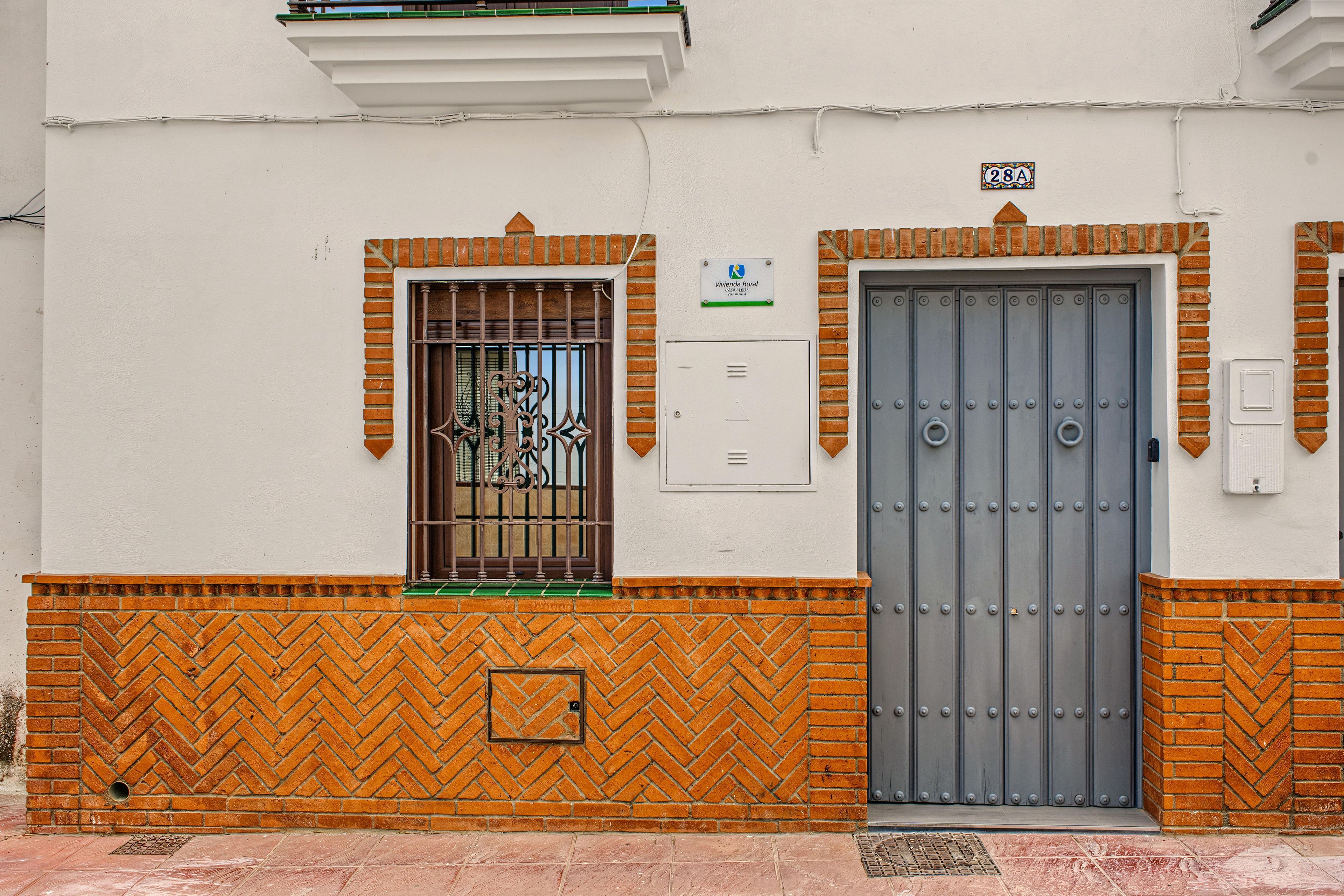 Fachada de edificio con puerta metálica, ventana con celosías decorativas y un cartel con dirección rural.