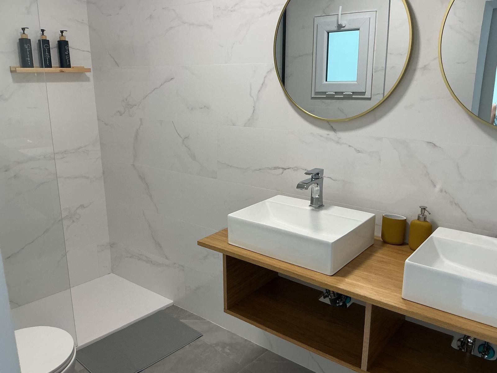 Un baño moderno con pared de mármol blanco, un lavamanos rectangular con un solo grifo, un inodoro y un espejo con marco dorado.