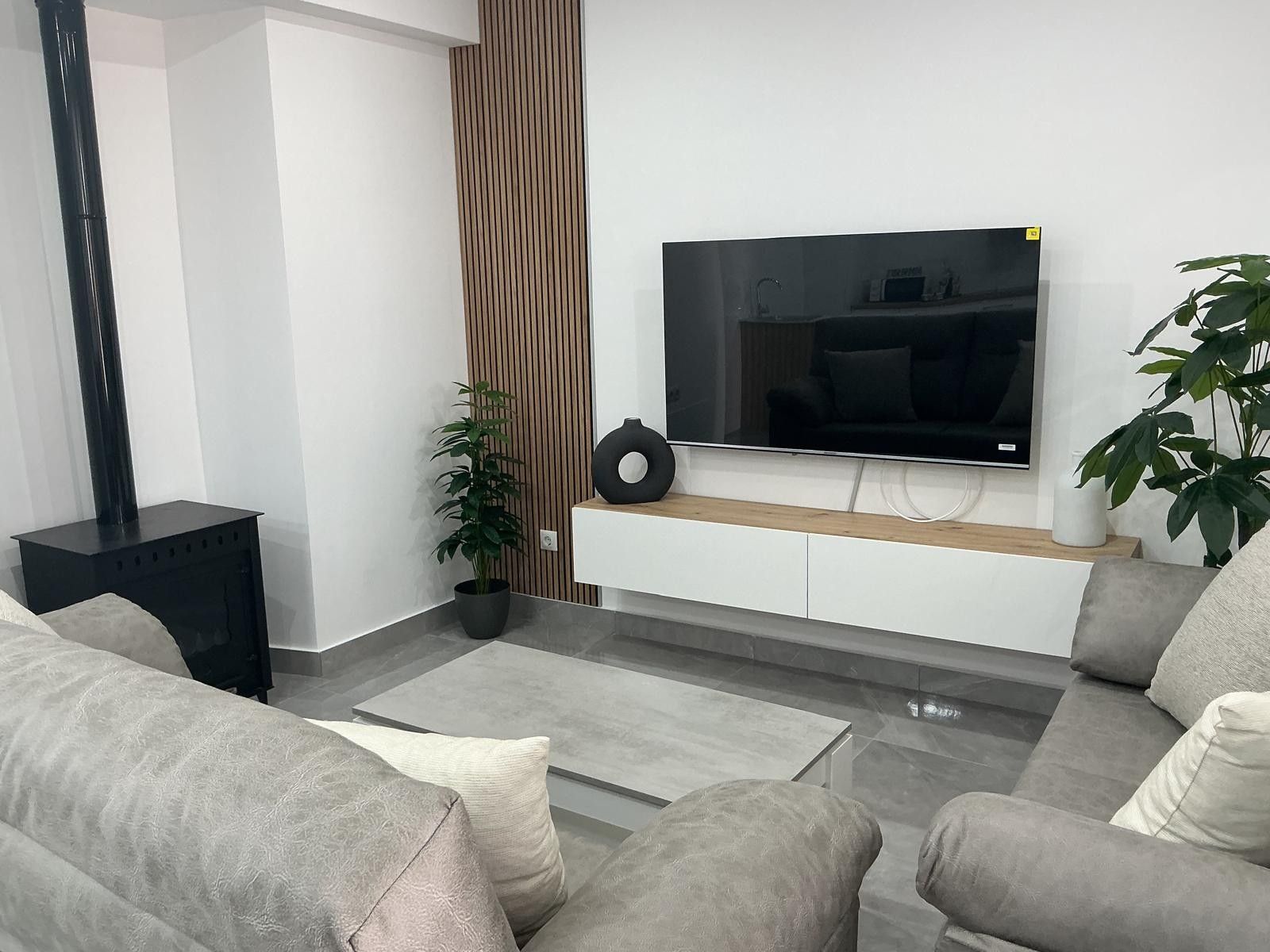 Un salón moderno con una televisión de pantalla plana montada en un mueble blanco, sofás grises, una mesita de noche negra y una planta en maceta.