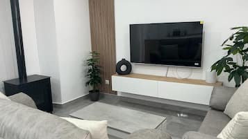 Un salón moderno con una televisión de pantalla plana montada en un mueble blanco, sofás grises, una mesita de noche negra y una planta en maceta.